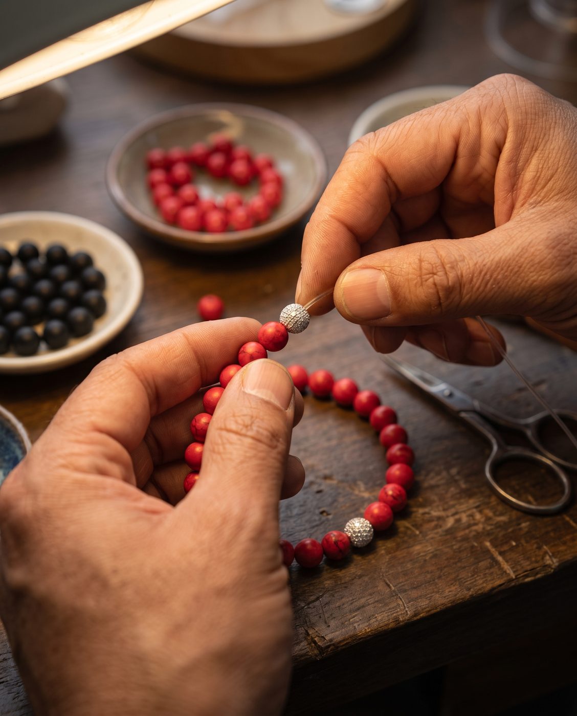 Hands threading red howlite beads onto a stretch cord at a workbench